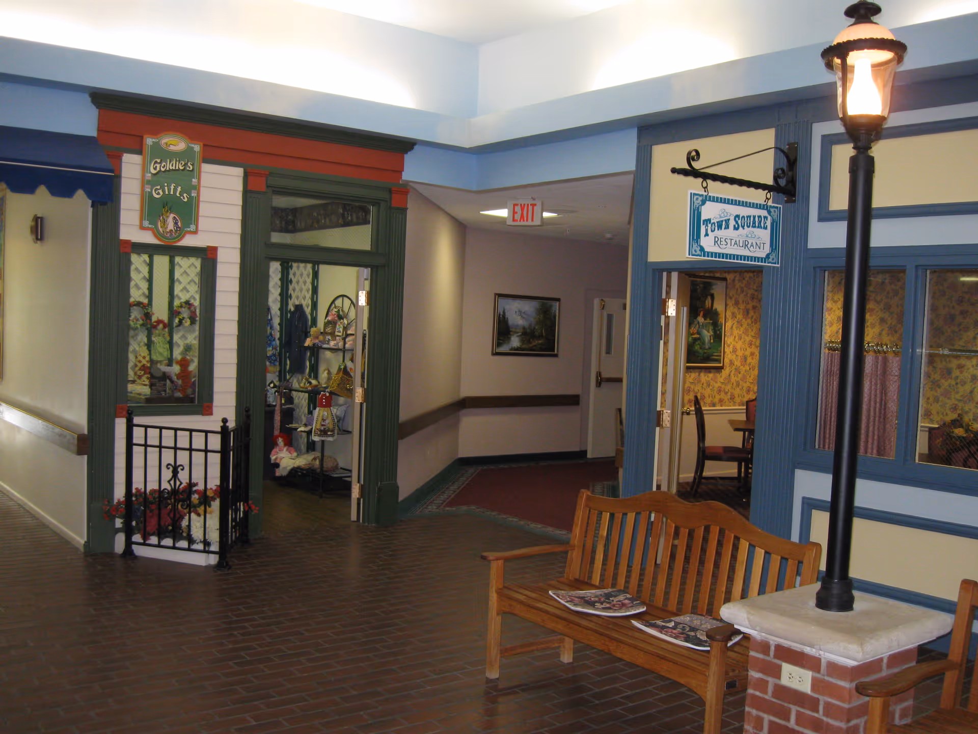 Indoor hallway of Seminary Village with storefront-style shops including 'Goldie's Gifts' and 'Town Square Restaurant', a wooden bench and a lamp post.