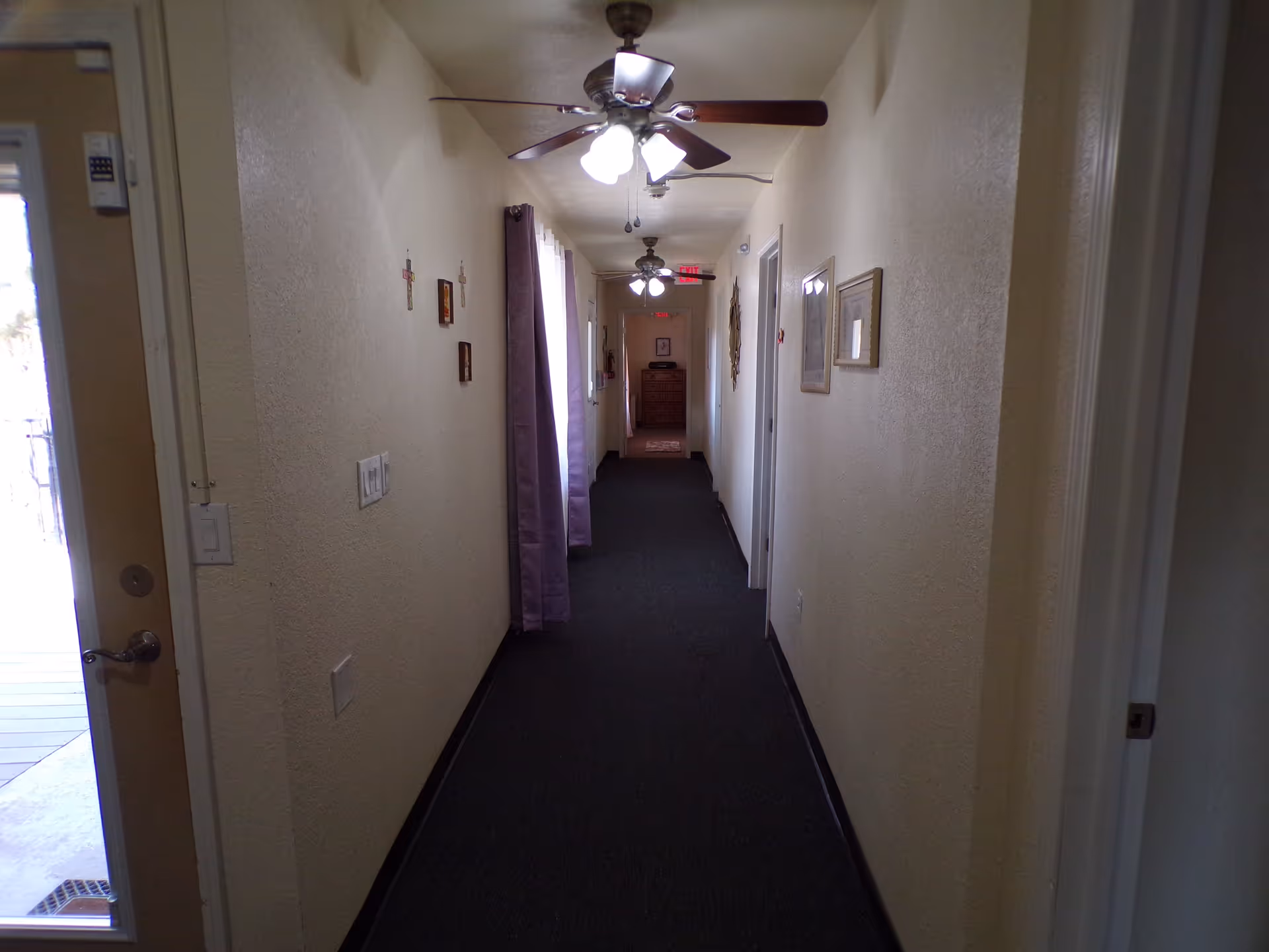 A long indoor hallway with beige walls and dark carpet. Ceiling fans with lights are mounted on the ceiling. There are framed pictures and decorative crosses on the walls. A door with a window is open on the left side near the front, letting in natural light. At the end of the hallway, there is a chest of drawers and an exit sign above a door.