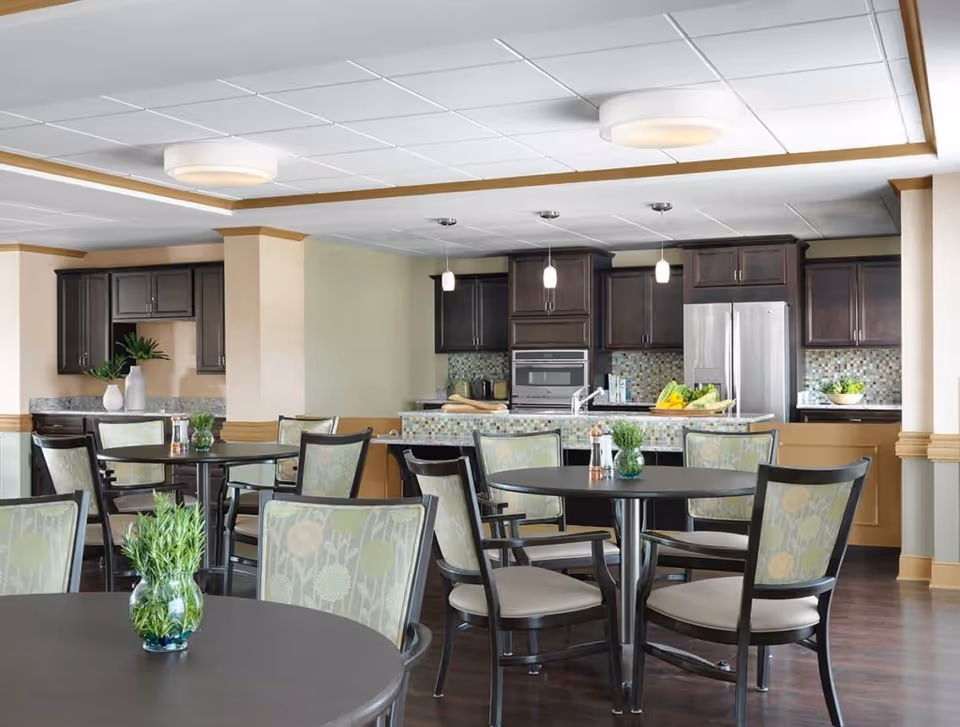 A bright and modern dining area with round tables and chairs featuring floral patterned upholstery. In the background, there is a kitchen with dark wood cabinets, a stainless steel refrigerator, built-in oven, and a mosaic tile backsplash. Small green plants in glass vases are placed on the tables, adding a fresh touch to the space.
