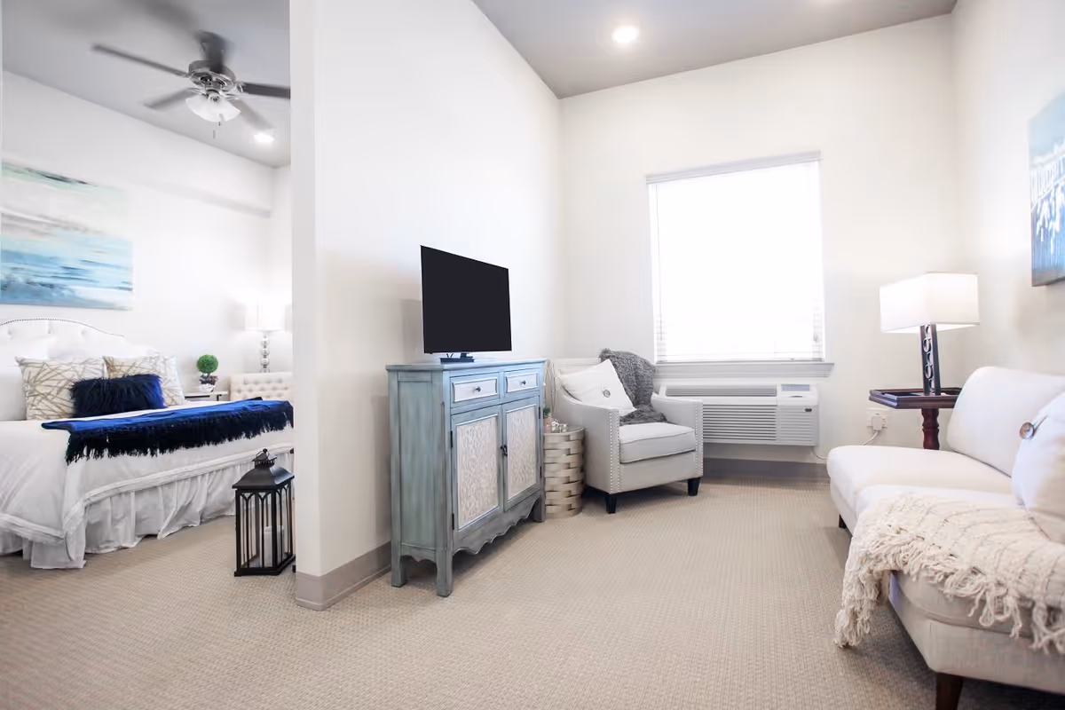A bright and airy living space featuring a small living room area with a white sofa, an armchair, a side table with a lamp, and a window with blinds. Adjacent to the living room is a bedroom with a bed adorned with white bedding, decorative pillows, a blue throw blanket, a nightstand with a lamp, and a ceiling fan above. A blue cabinet with a flat-screen TV sits between the living room and bedroom areas.