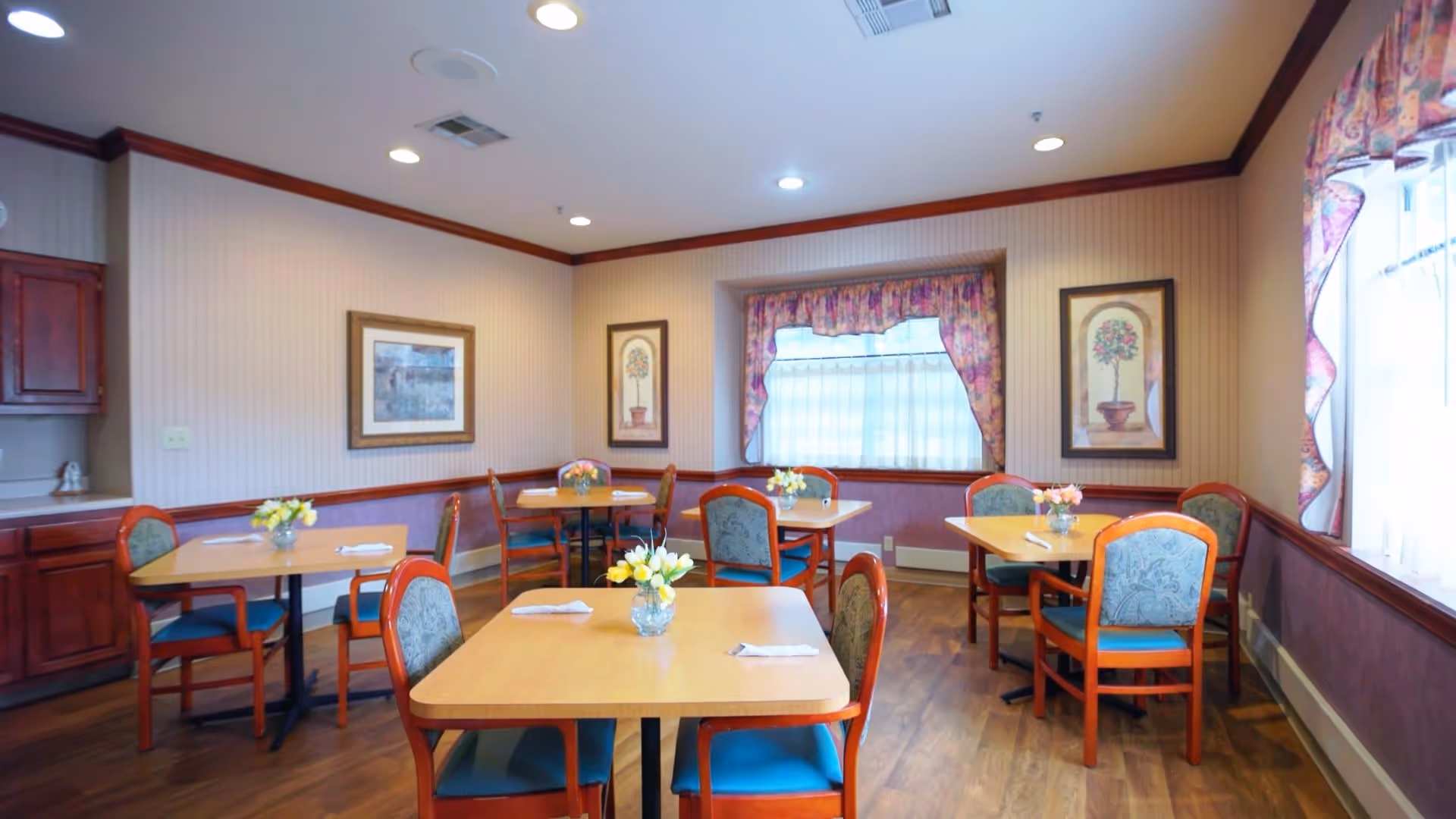 A cozy dining room with several wooden tables and chairs arranged neatly. Each table has a small vase with flowers and napkins placed on it. The room features wood flooring, soft lighting from ceiling fixtures, framed artwork on the walls, and windows with floral curtains allowing natural light to enter.