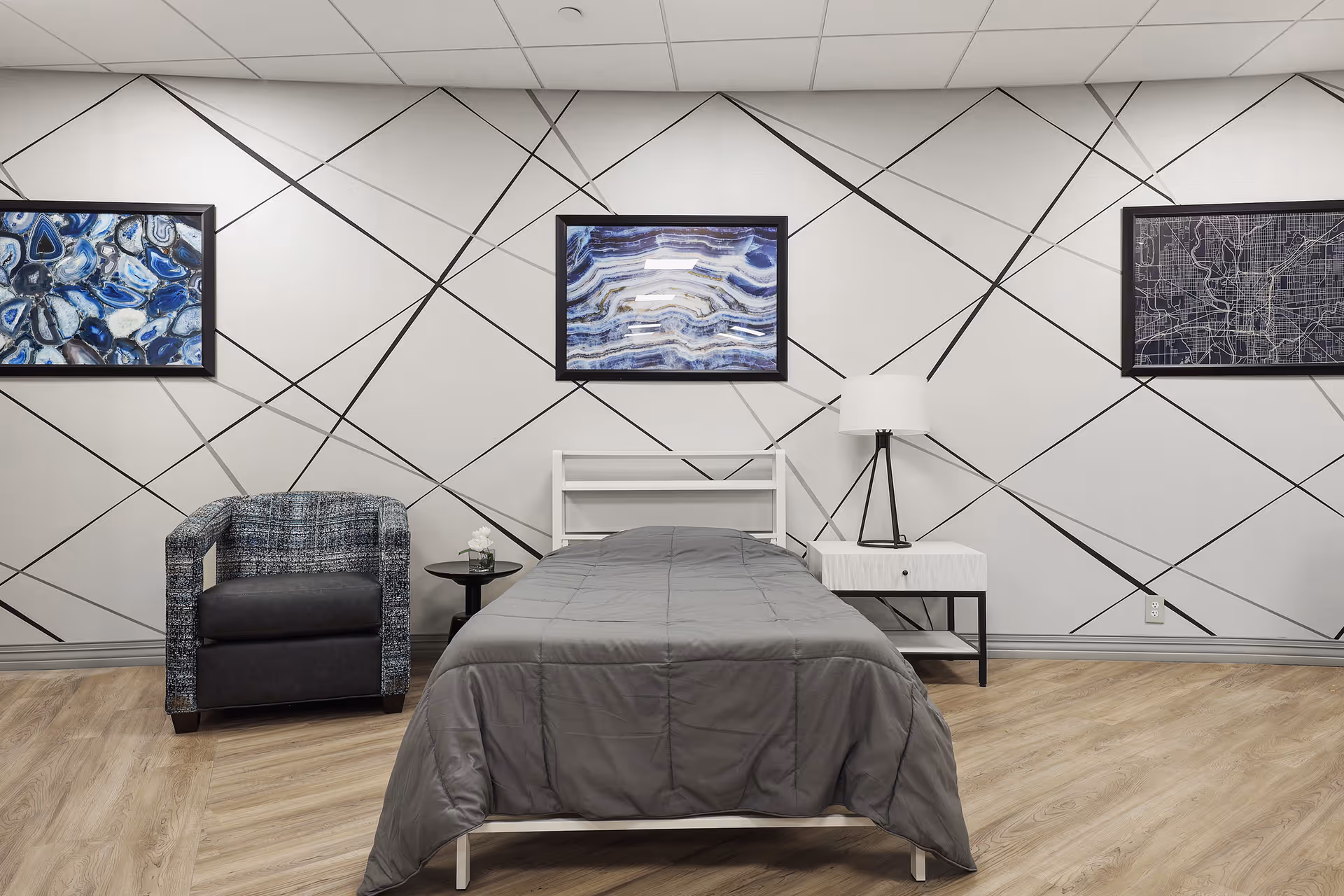 A modern bedroom with a single bed covered in a gray comforter, a white nightstand with a lamp, a small round side table with a flower vase, and a patterned armchair. The wall behind features a geometric design with diagonal black lines and three framed artworks.