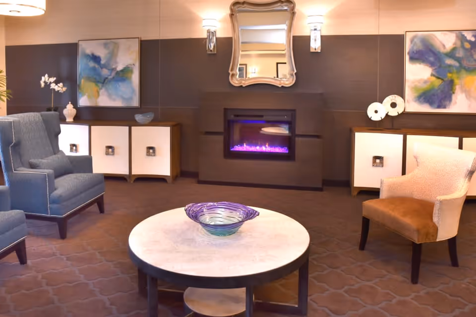 A cozy living room area with a round marble coffee table holding a decorative glass bowl in the center. There are two upholstered armchairs, one blue and one beige with a brown seat, positioned around the table. The back wall features a modern electric fireplace with purple flames, a decorative mirror above it, and two abstract paintings on either side. The room has warm lighting and a patterned carpet.