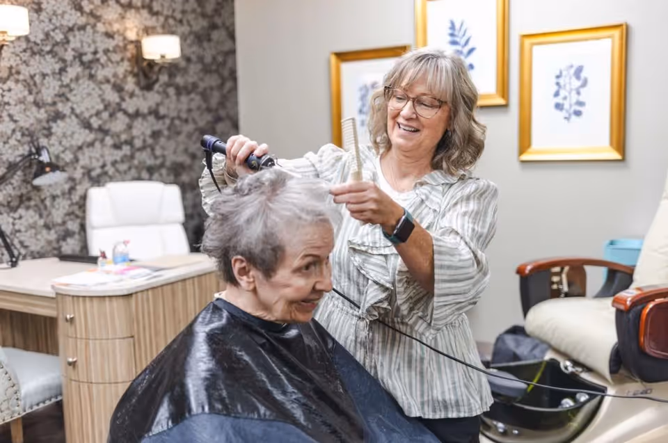 A senior woman is getting her hair styled by a smiling hairstylist in a cozy room with floral wallpaper, framed botanical prints on the wall, and comfortable seating.