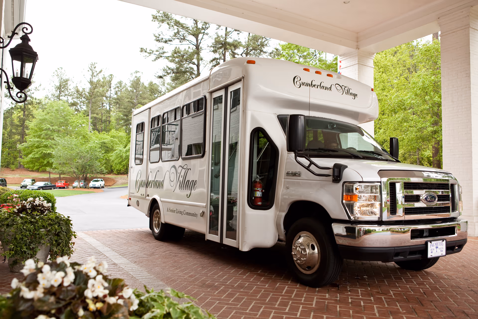 A white shuttle bus parked under a covered entrance with the name Cumberland Village written on the side and front. The bus is surrounded by greenery and flowers with a brick-paved driveway.