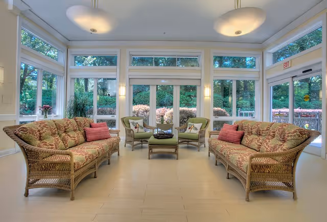 A bright and spacious living room area with large windows showing a green outdoor garden. The room features two wicker sofas with floral cushions and pink throw pillows, two wicker armchairs with green cushions, and a matching wicker ottoman in the center. Two ceiling lights hang above, and there are wall sconces on either side of the central window. The floor is light-colored wood or tile.