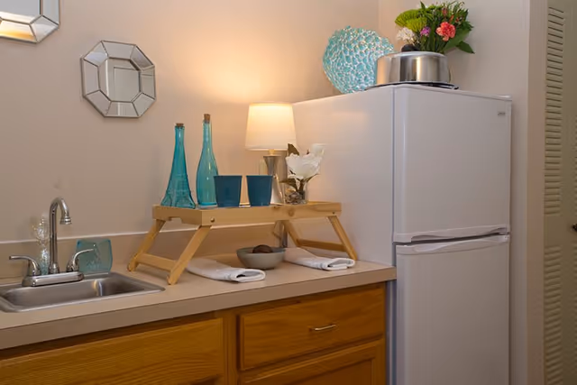 A small kitchen area with a white refrigerator, wooden cabinets, and a countertop with a sink. On the countertop, there is a wooden tray holding two blue glasses and two blue bottles, a small lamp, a white flower in a vase, and a bowl with some items. Decorative mirrors hang on the wall above the sink.