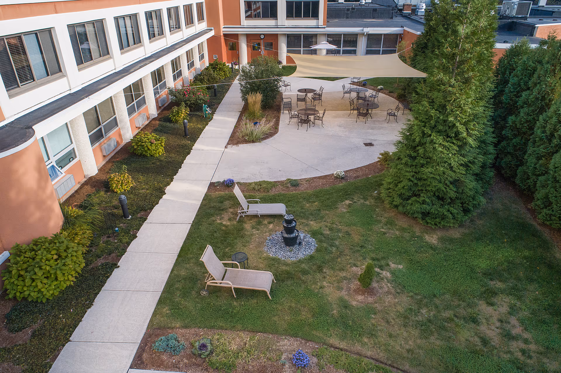 Outdoor courtyard area at Ludlowe Center for Health and Rehabilitation featuring a paved walkway, several patio tables with chairs under a large shade sail, two lounge chairs near a small water fountain, surrounded by greenery and trees.