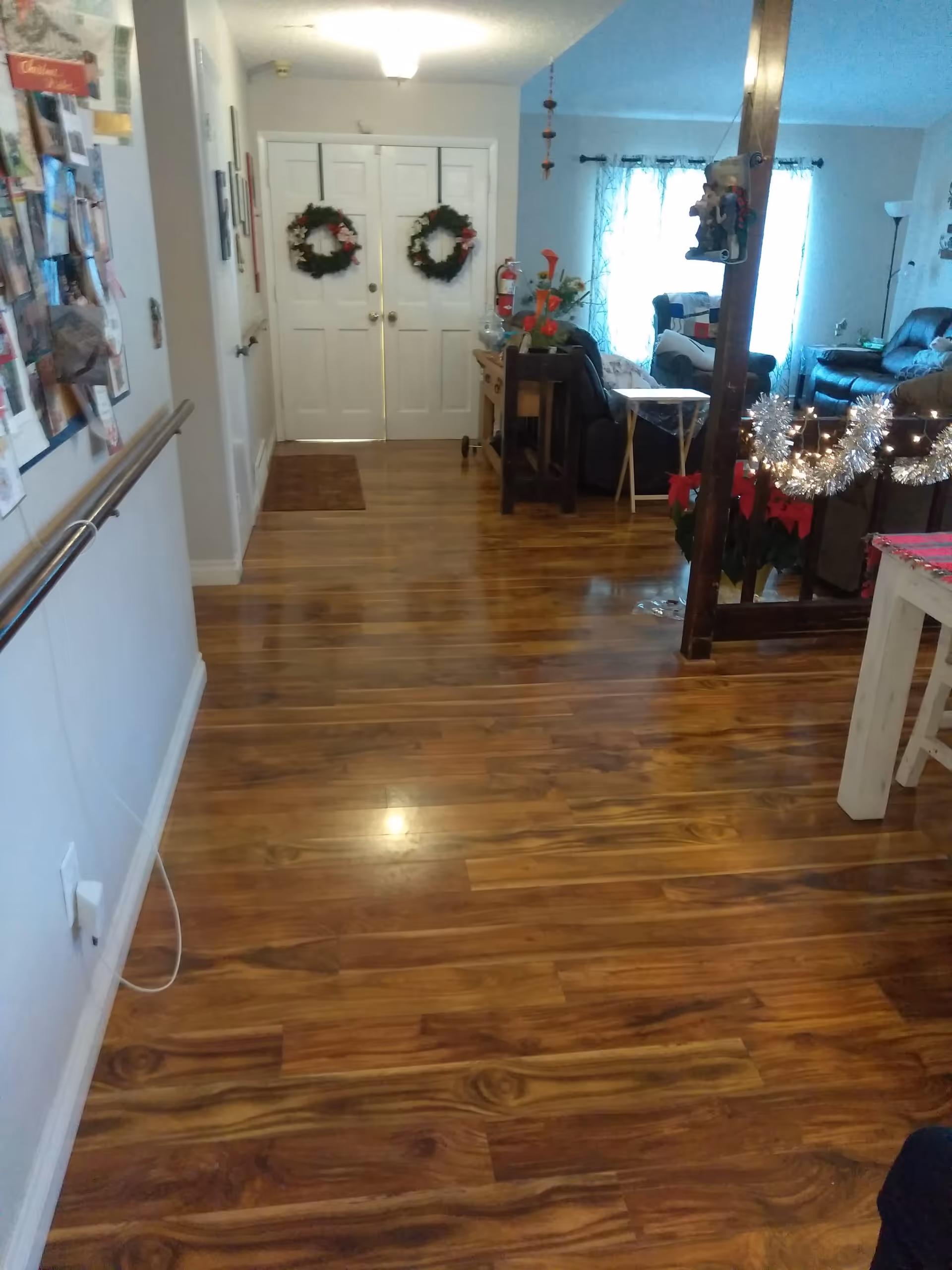 Interior view of a senior living facility hallway with wooden flooring leading to double white doors decorated with Christmas wreaths. To the right, there is a living area with chairs, a couch, and holiday decorations including tinsel and poinsettias. A bulletin board with various papers is visible on the left wall.