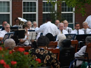 A group of musicians playing instruments outdoors in front of a brick building with large windows, with an audience seated and watching the performance.