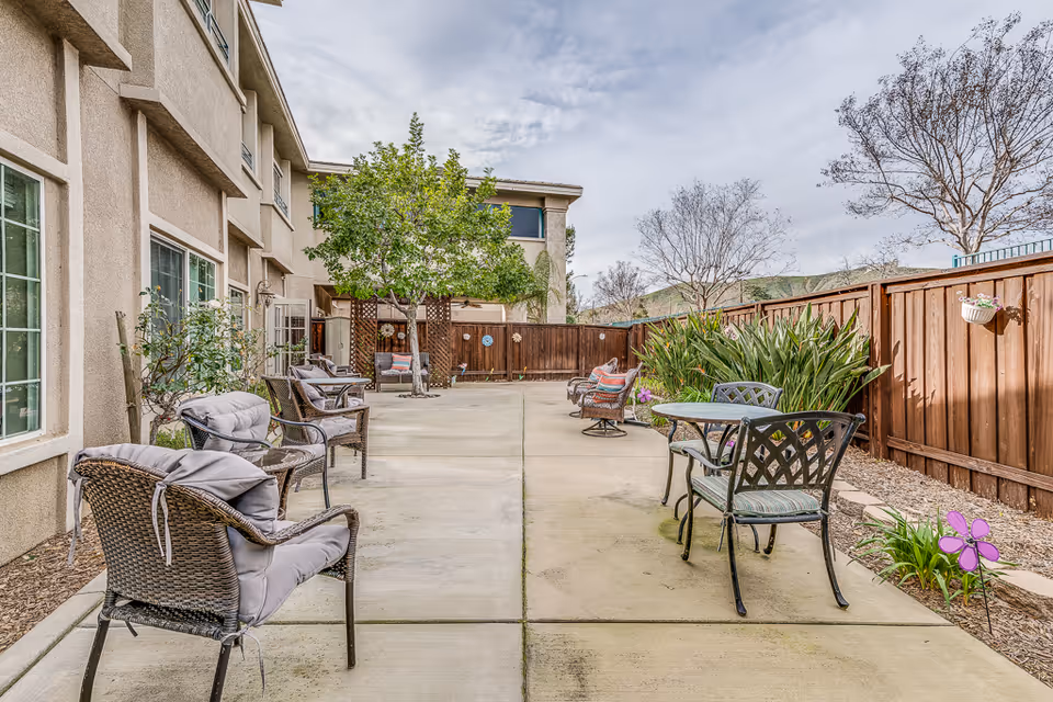 Outdoor patio area at Villas at San Bernardino with cushioned wicker chairs and metal chairs around small tables, surrounded by plants and a wooden fence under a cloudy sky.