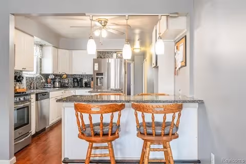 Bright kitchen with a granite breakfast bar, two wooden bar stools, stainless steel appliances, and pendant lighting.