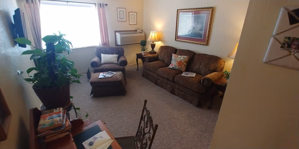 A cozy living room with a brown patterned sofa and matching armchair with ottoman. There are two side tables with lamps, a framed picture on the wall, a large window with curtains, a potted plant on a stand, and a small desk with books and papers.