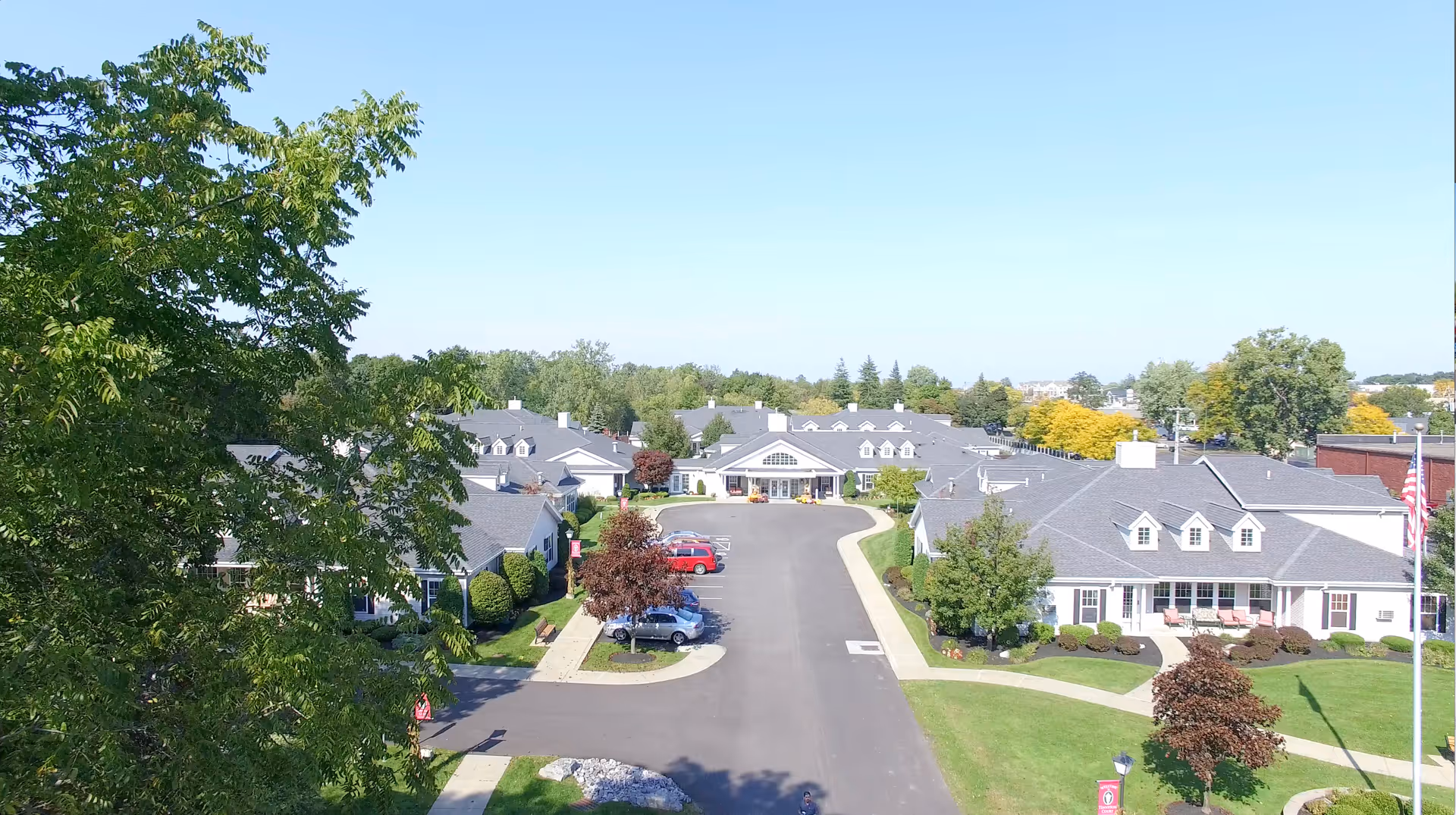 Aerial view of Tennyson Court senior living facility showing multiple single-story buildings with gray roofs, surrounded by green lawns, trees, and a paved driveway with parked cars under a clear blue sky.