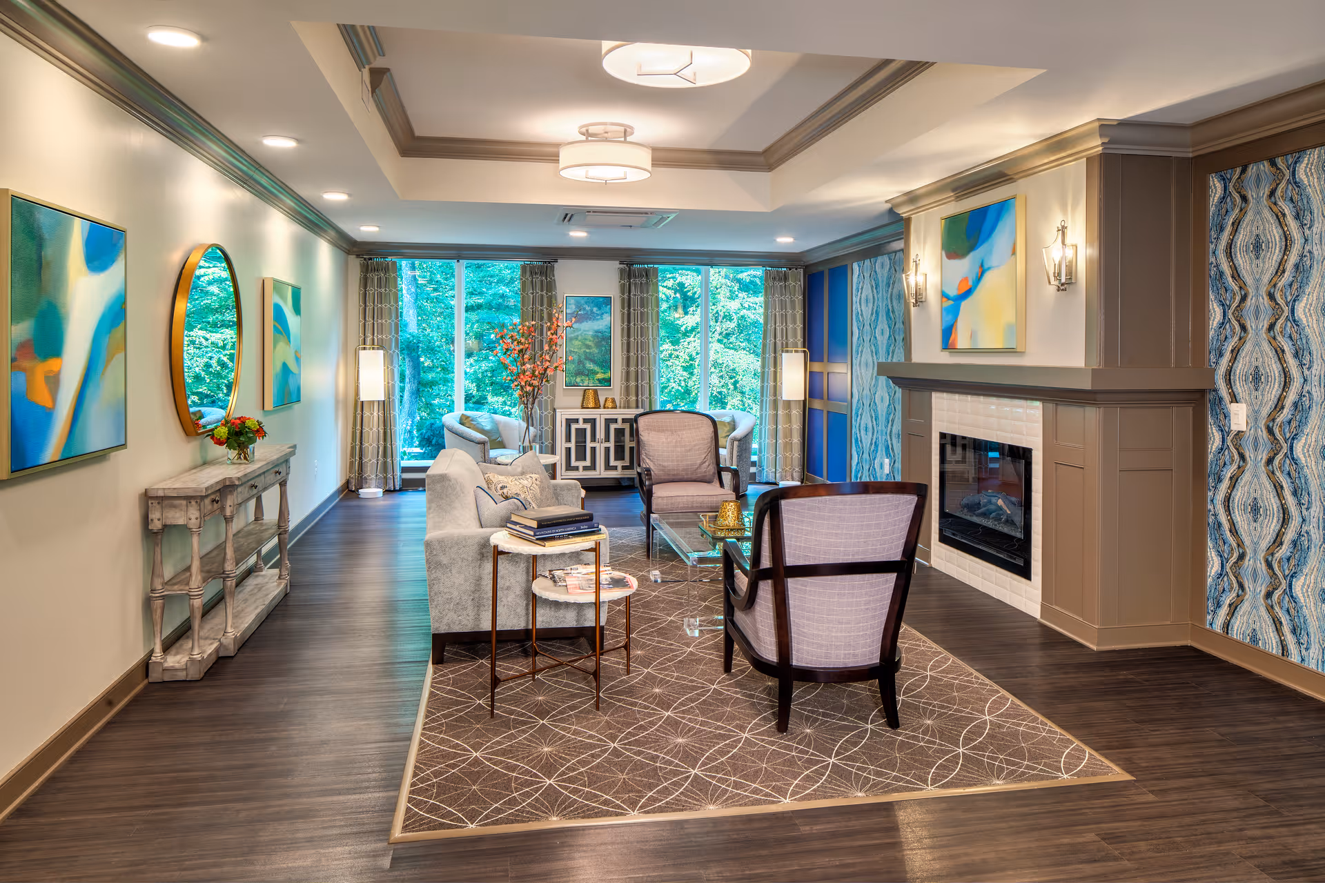A modern living room with a fireplace, two armchairs, a sofa, a glass coffee table, and a patterned area rug. The room features large windows with curtains, abstract artwork on the walls, and a console table with a round mirror and flowers. The walls have decorative paneling and wallpaper accents.