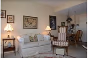 A cozy living room featuring a white sofa with patterned cushions, a striped armchair with an ottoman, two side tables each with a lamp, framed artwork on the walls, and a dining area with a wooden table and chairs in the background.
