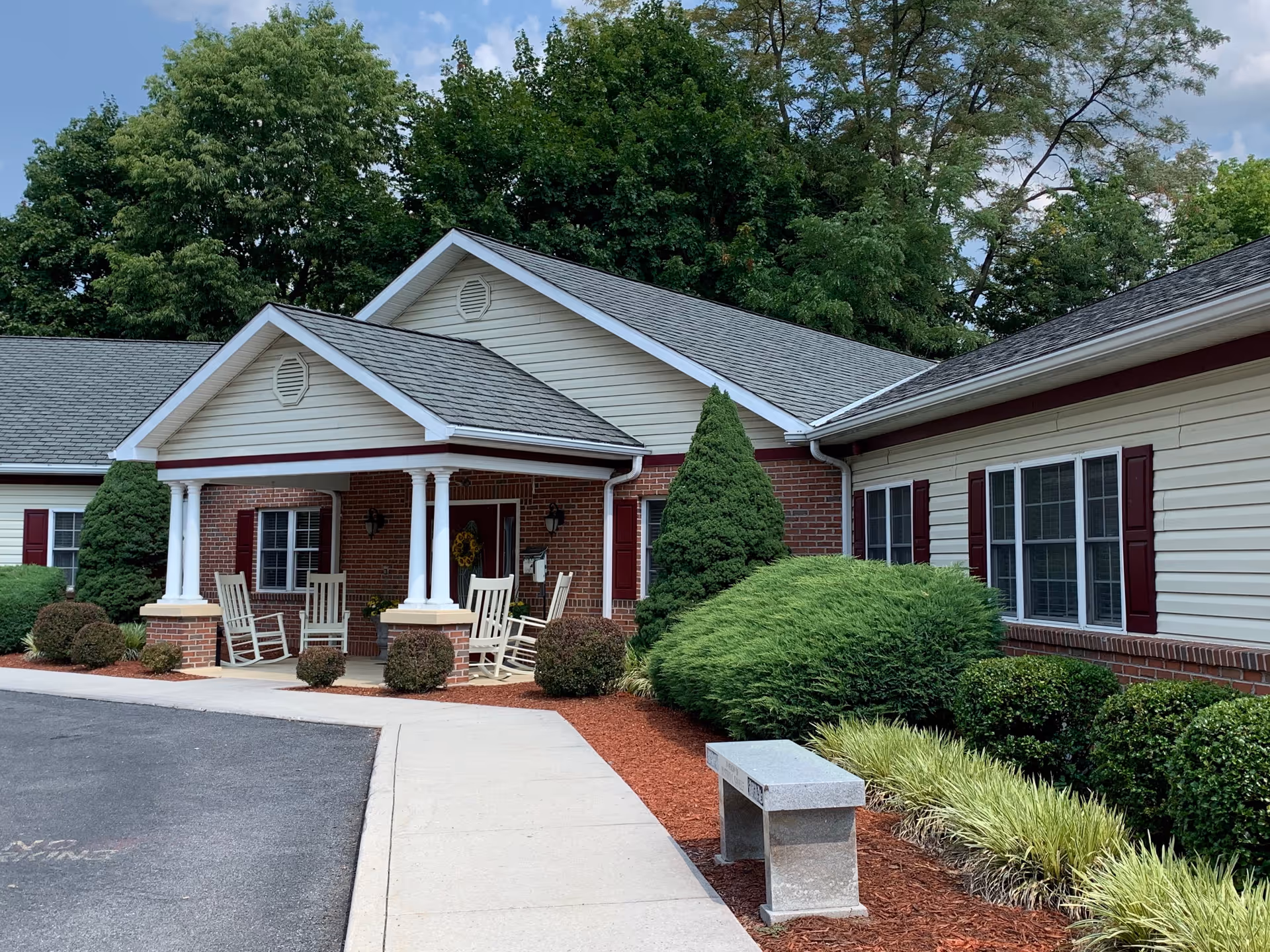 Exterior view of a single-story residential building with a covered porch featuring white columns and rocking chairs. The building has a combination of brick and light-colored siding with maroon shutters. There are well-maintained shrubs and bushes along the walkway leading to the entrance, and a stone bench is placed near the path. Trees and a partly cloudy sky are visible in the background.