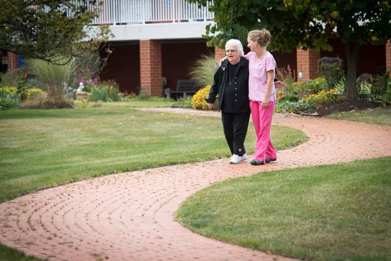 An elderly woman and a younger woman walking arm in arm along a curved brick pathway in a garden area with green grass, trees, and flowering plants near a building.