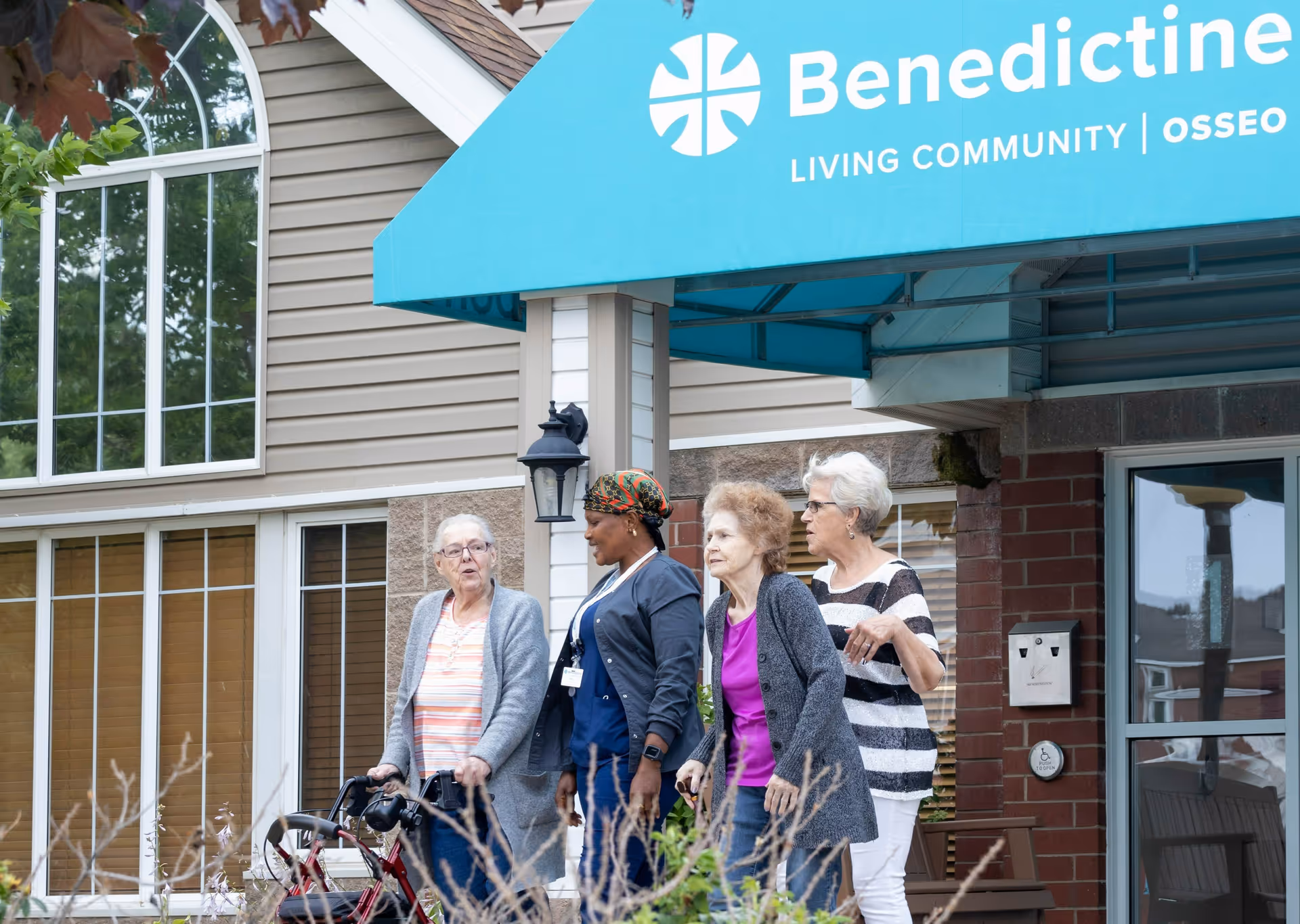 Four elderly women standing and walking outside the entrance of Benedictine Living Community in Osseo. One woman is using a walker, and the others are engaged in conversation near the building's entrance with a blue awning displaying the facility's name.