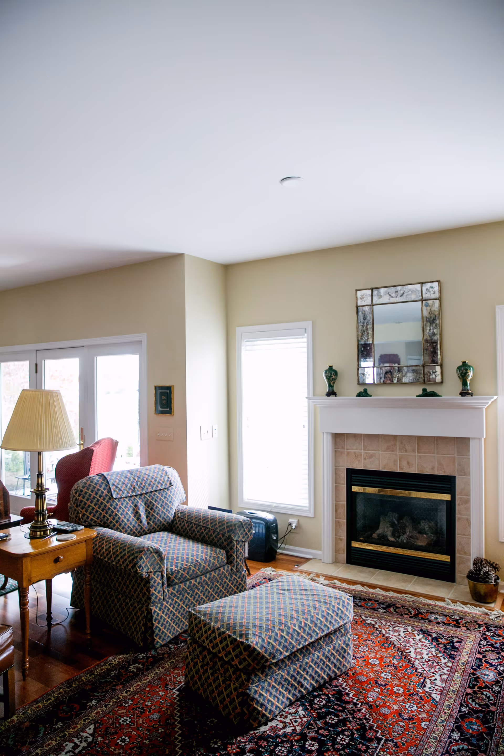 A cozy living room with a patterned armchair and matching ottoman placed on a red and blue patterned rug. There is a wooden side table with a lamp and remote controls next to the armchair. The room features a fireplace with a tiled surround and a decorative mirror above it, flanked by two green vases. Two windows with white blinds allow natural light into the space, and glass doors lead outside.