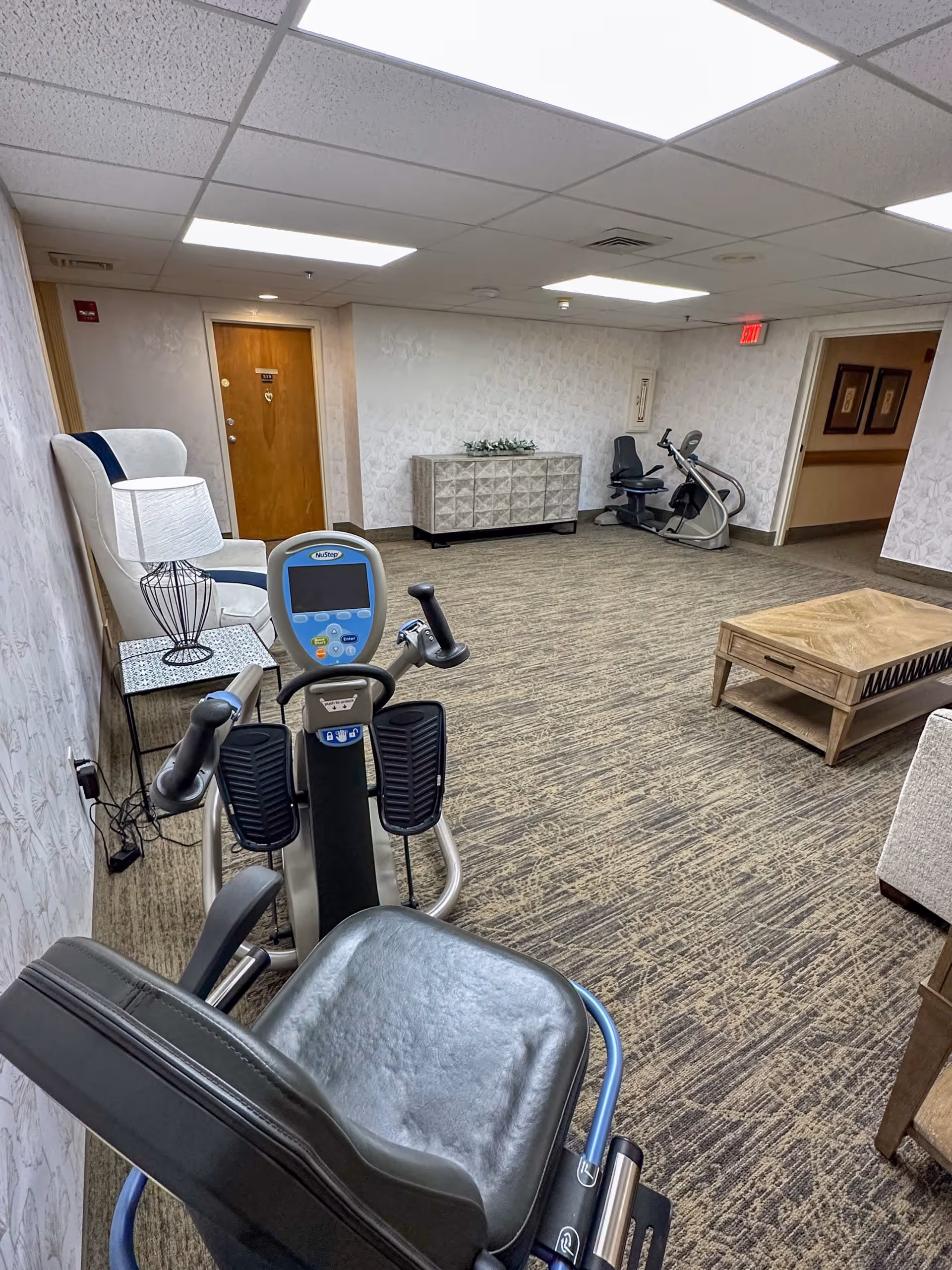 Interior room in a senior living facility with exercise equipment including a NuStep recumbent cross trainer and another stationary bike. The room has patterned carpet, wallpapered walls, a wooden door, a side table with a lamp, a wooden coffee table, and a decorative cabinet against the wall.