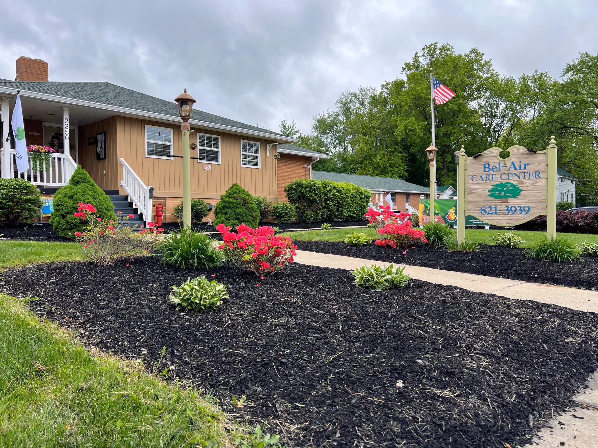 Exterior view of Bel-Air Care Center, a single-story building with beige siding and white trim. There is a small porch with white railings and steps leading up to the entrance. The landscaped front yard features neatly trimmed bushes, vibrant pink flowers, and a mulched garden bed. An American flag flies on a pole next to a wooden sign displaying the facility's name and phone number. The sky is overcast and trees are visible in the background.