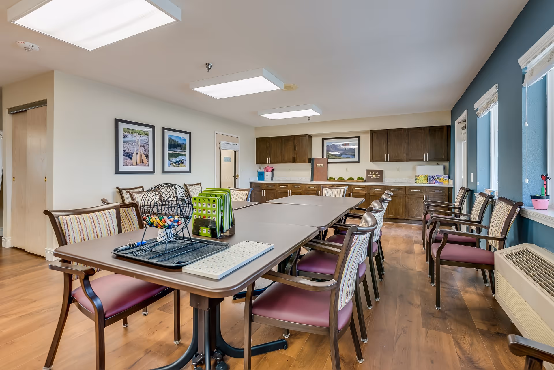 A well-lit activity room with a long table surrounded by chairs. On the table, there is a bingo cage with colorful balls and bingo cards. The room has wooden flooring, light-colored walls, and several framed pictures on the wall. There are windows with blinds on the right side and wooden cabinets with a countertop at the back.