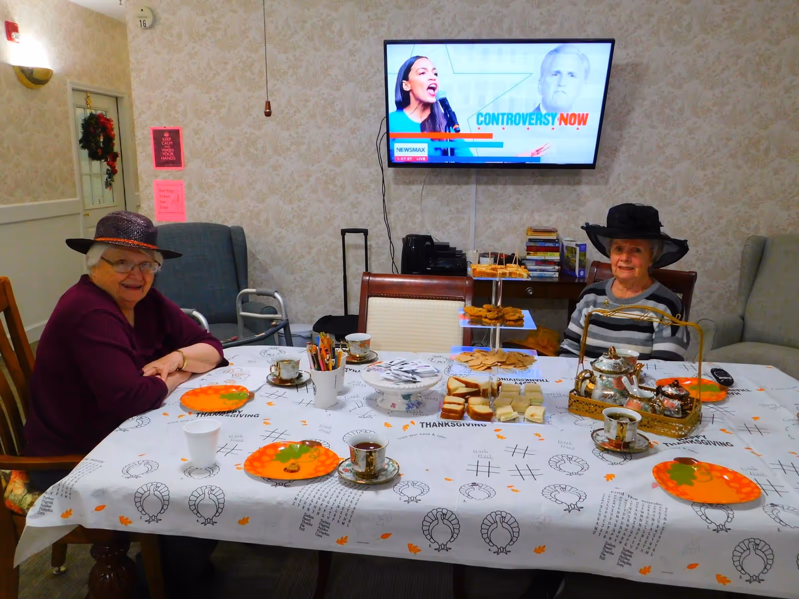 Two elderly women wearing hats sit at a table set with tea, plates and snacks on a Thanksgiving-themed tablecloth in a communal room with a TV on the wall.
