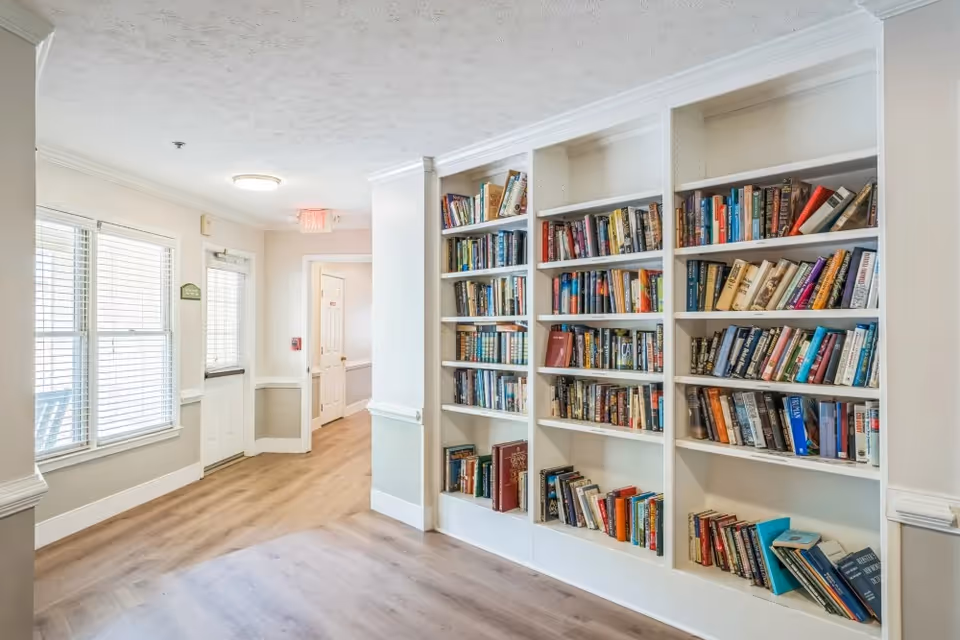 A bright hallway in a senior living facility with light wood flooring, white walls, and a built-in bookshelf filled with various books. There is a door with a window and blinds on the left side, and a hallway leading to other rooms in the background.