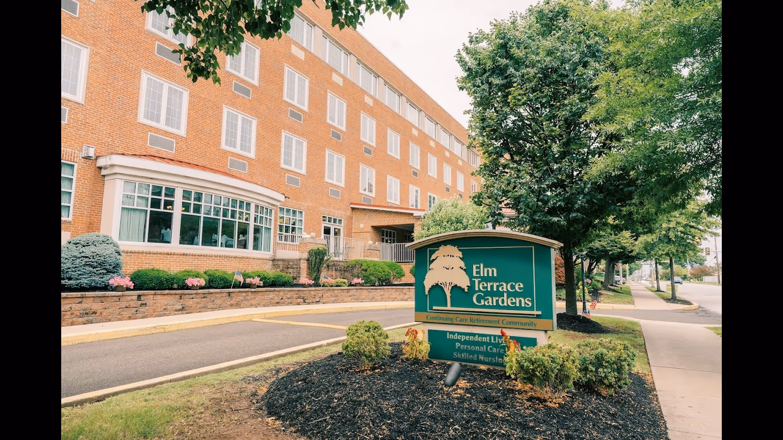 Brick senior living building with landscaped entrance and a green sign reading "Elm Terrace Gardens".