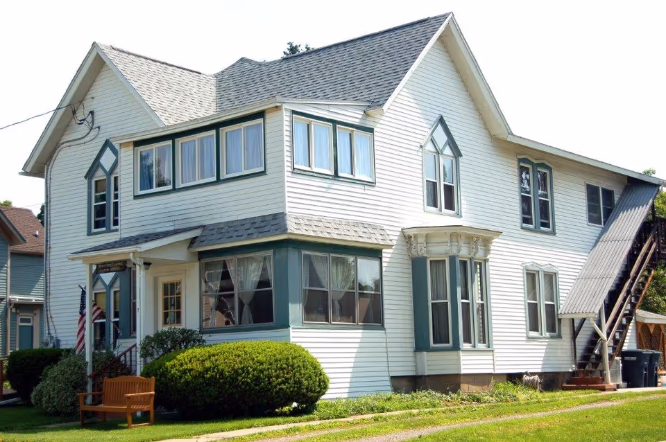 White two-story residential building with green-trimmed windows, a small front porch, shrubs, a bench, and an exterior staircase.