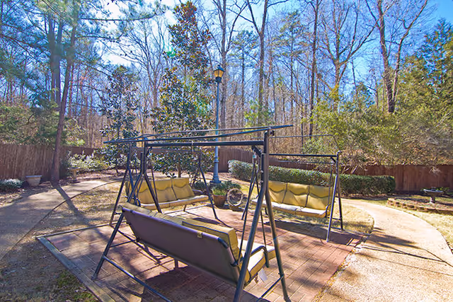Outdoor seating area with three metal-framed swings with yellow cushions arranged around a paved patio in a garden setting. The area is surrounded by trees, bushes, and a wooden fence under a clear blue sky.