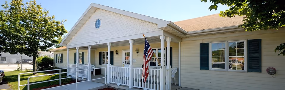 Exterior view of a single-story building with beige siding and a covered porch supported by white columns. The porch has a white railing and an American flag displayed. There are green shutters on the windows and a tree on the left side of the building. The building number 1016 is visible above the porch.