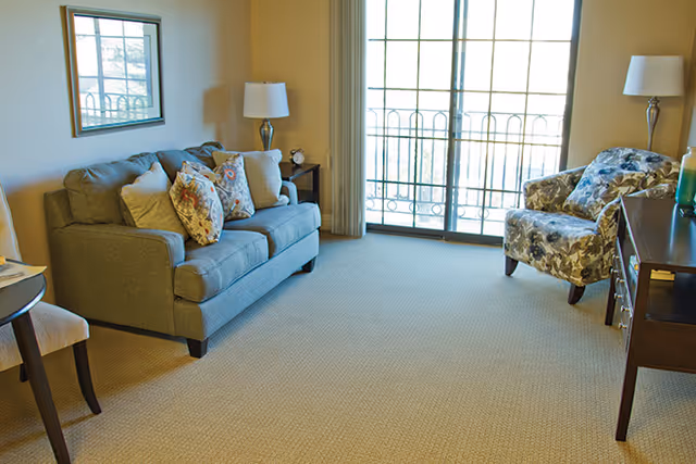 A cozy living room with a gray sofa adorned with patterned cushions, a floral armchair, a wooden side table with a lamp, and a large window with vertical blinds letting in natural light.
