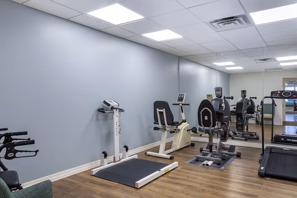 A small fitness room with exercise equipment including a treadmill, stationary bikes, and a weight machine. The room has light blue walls, wood flooring, and a large mirror covering one wall.