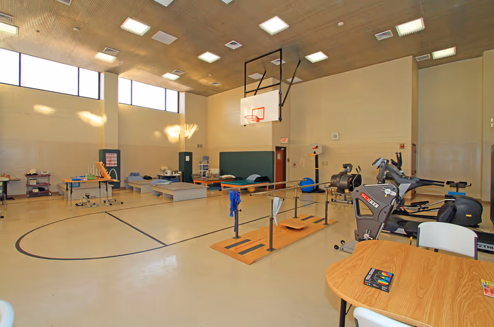 A large indoor physical therapy or rehabilitation room with exercise equipment, parallel bars for walking practice, therapy beds, and a basketball hoop mounted on the wall. The room has high windows letting in natural light and a wooden ceiling with multiple ceiling lights.