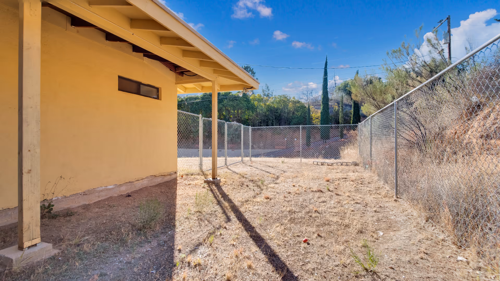 Outdoor fenced area next to a yellow building with a covered porch, dry grass, and some sparse vegetation under a blue sky with scattered clouds.