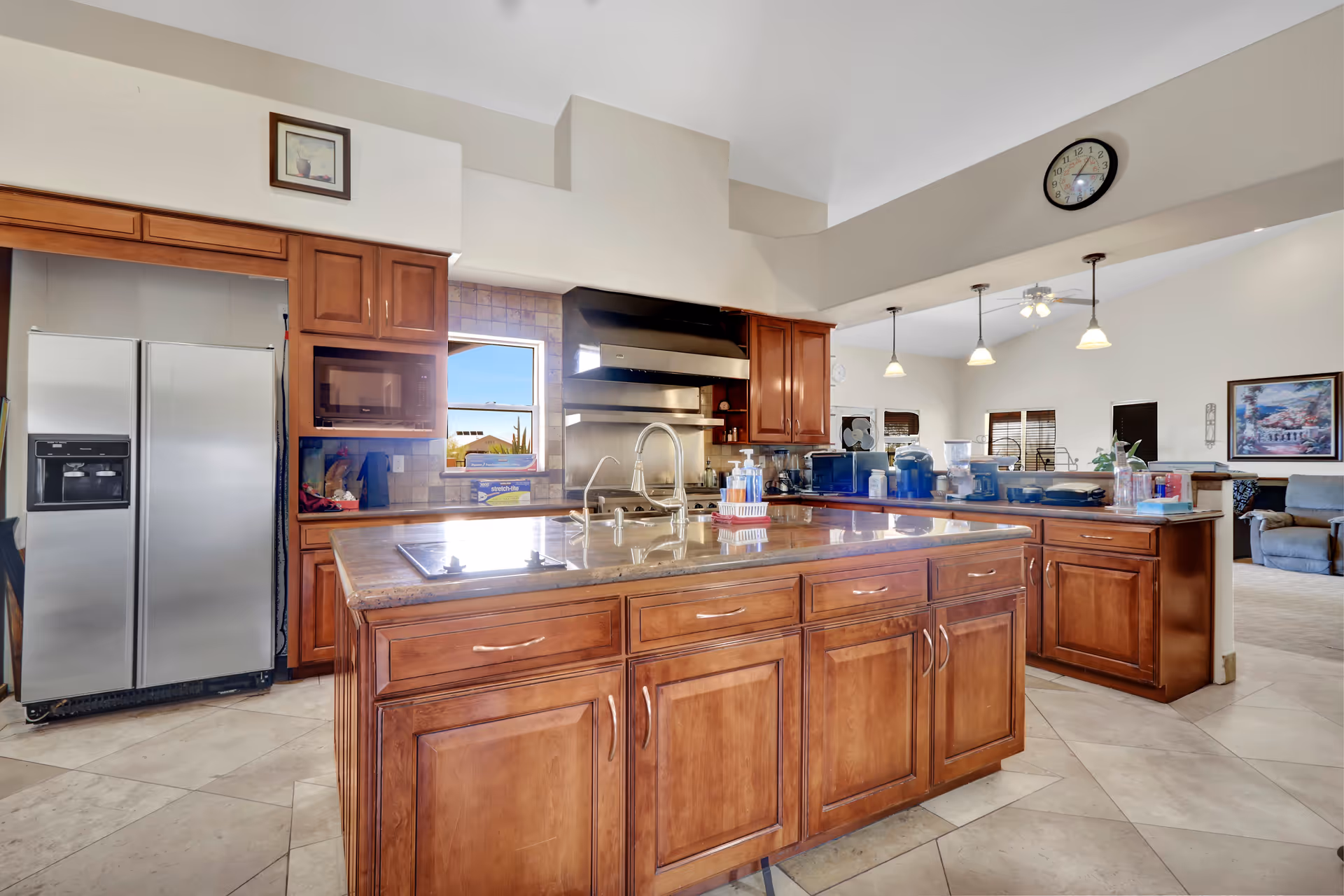 Spacious kitchen with a large wooden island, sink, stovetop, stainless steel refrigerator and cabinets, open to a living area.