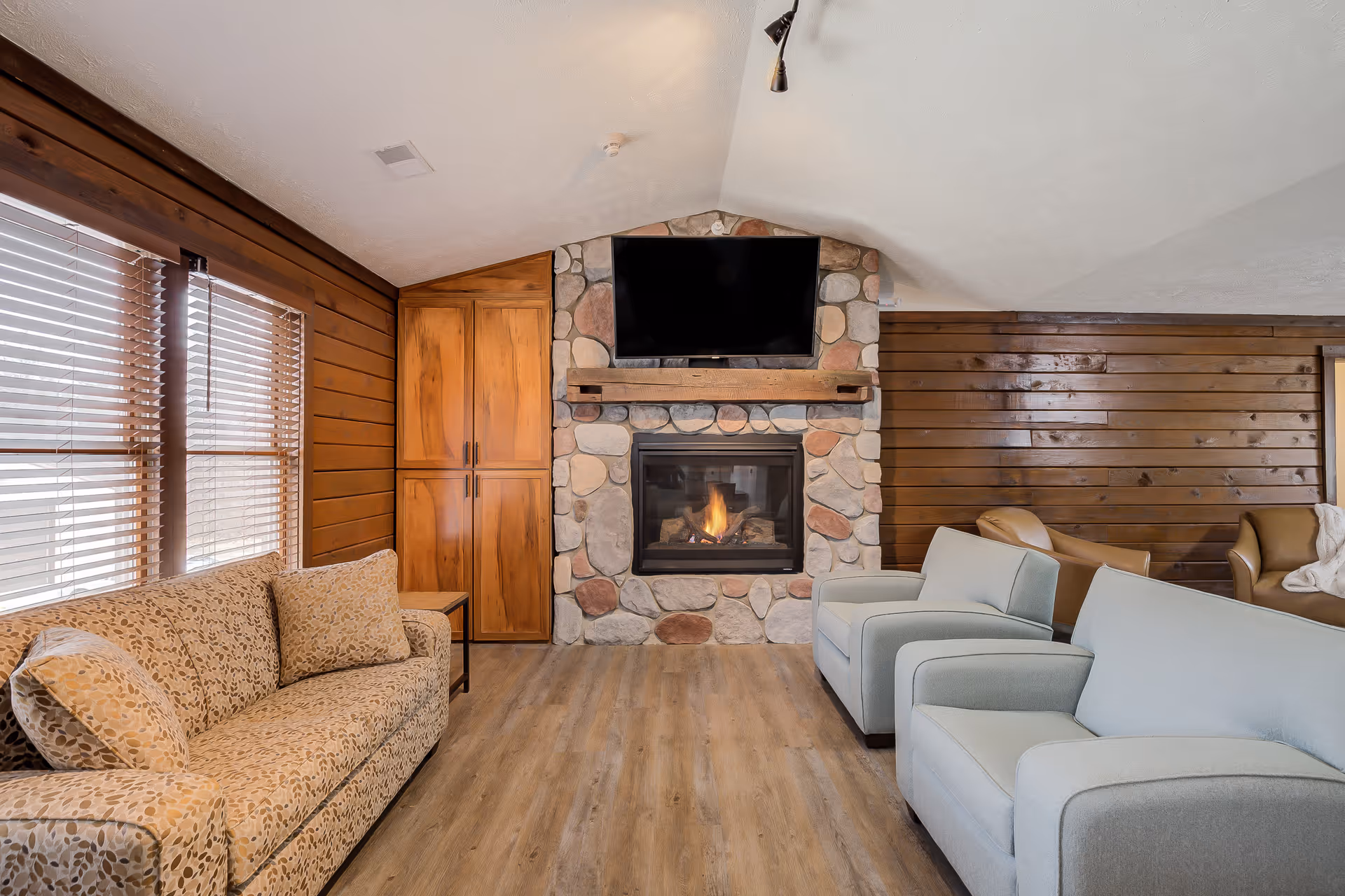 Cozy living room with a stone fireplace and a mounted flat-screen TV above it. The room features wooden walls, a patterned beige sofa with matching pillows on the left, two light blue armchairs in the center, and additional seating in the background. Large windows with wooden blinds allow natural light to enter the space.