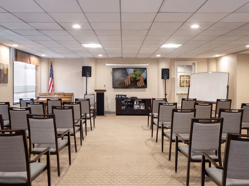 A meeting/activity room with rows of chairs facing a podium, wall-mounted TV, speakers, and a whiteboard.