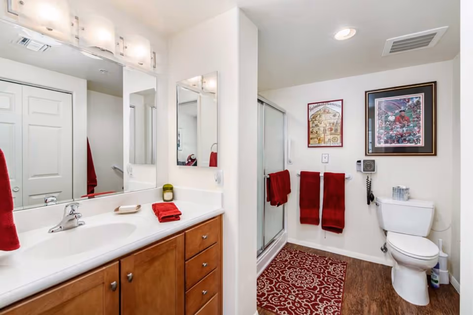 A clean and well-lit bathroom with a wooden vanity featuring a white countertop and sink. Above the sink is a large mirror with three light fixtures. To the right, there is a glass-enclosed shower with red towels hanging on a grab bar. A white toilet is next to the shower, with a telephone mounted on the wall above it. The floor has a red patterned rug, and two framed pictures hang on the wall above the toilet. The bathroom has white walls and a wood-look floor.