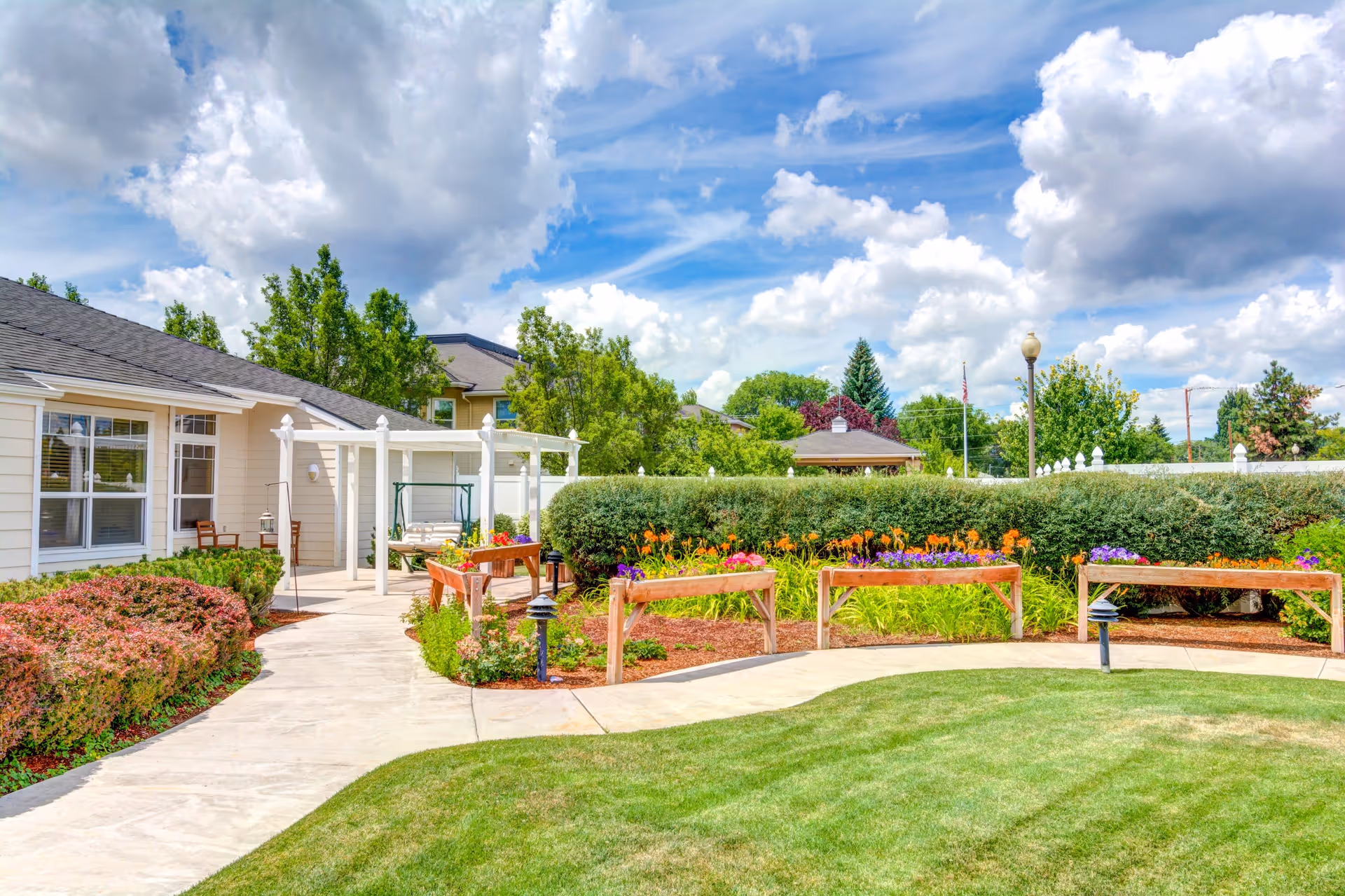 A bright outdoor garden area at Pelican Pointe by Cogir featuring a curved concrete walkway, green grass, colorful flower beds with orange and purple flowers, wooden raised garden beds, and a white pergola with a swing bench. The sky is partly cloudy with blue patches, and there are trees and shrubs surrounding the garden.