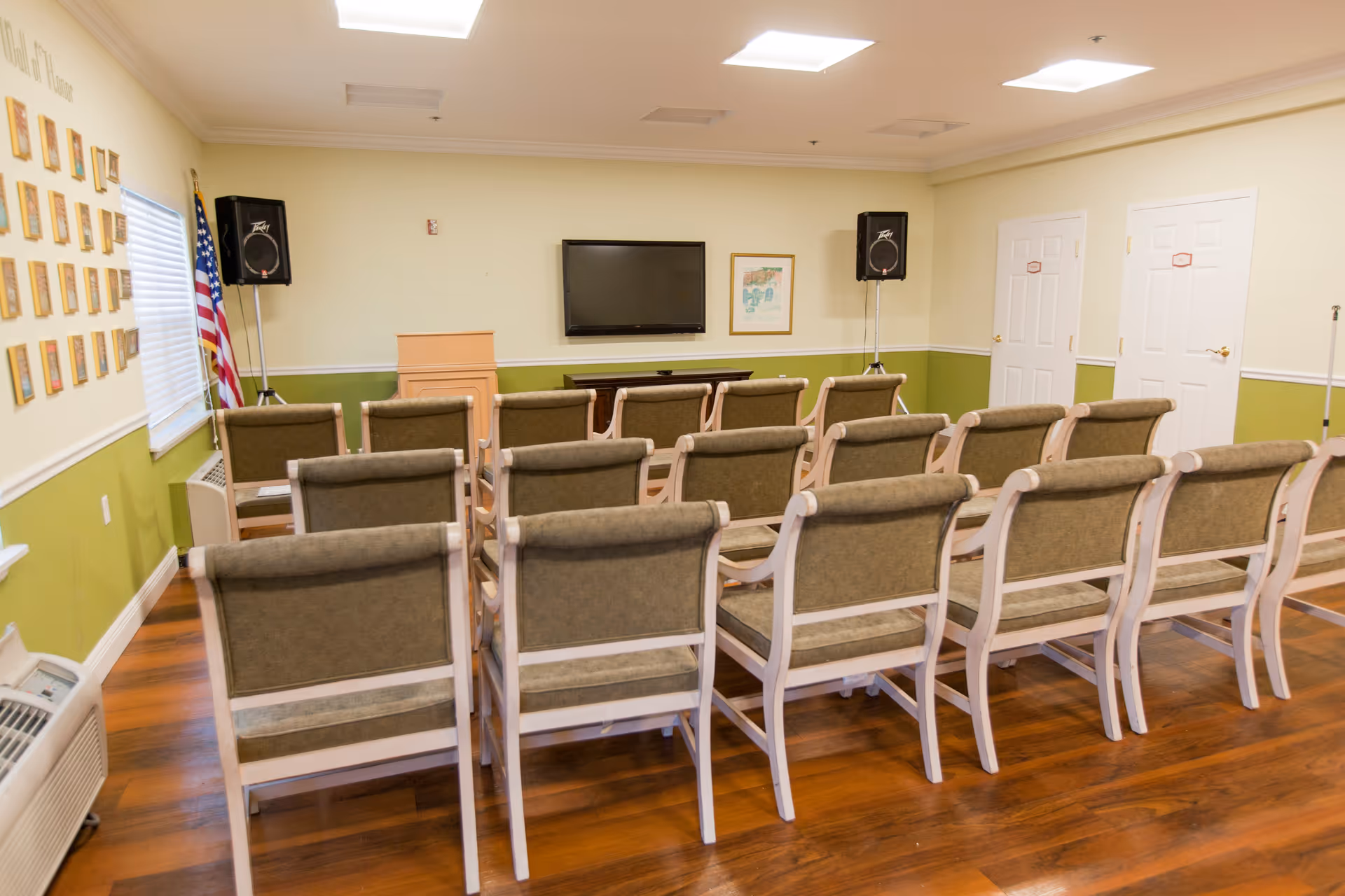 Rows of chairs facing a wall-mounted TV and podium in a small meeting/activity room with speakers and an American flag.