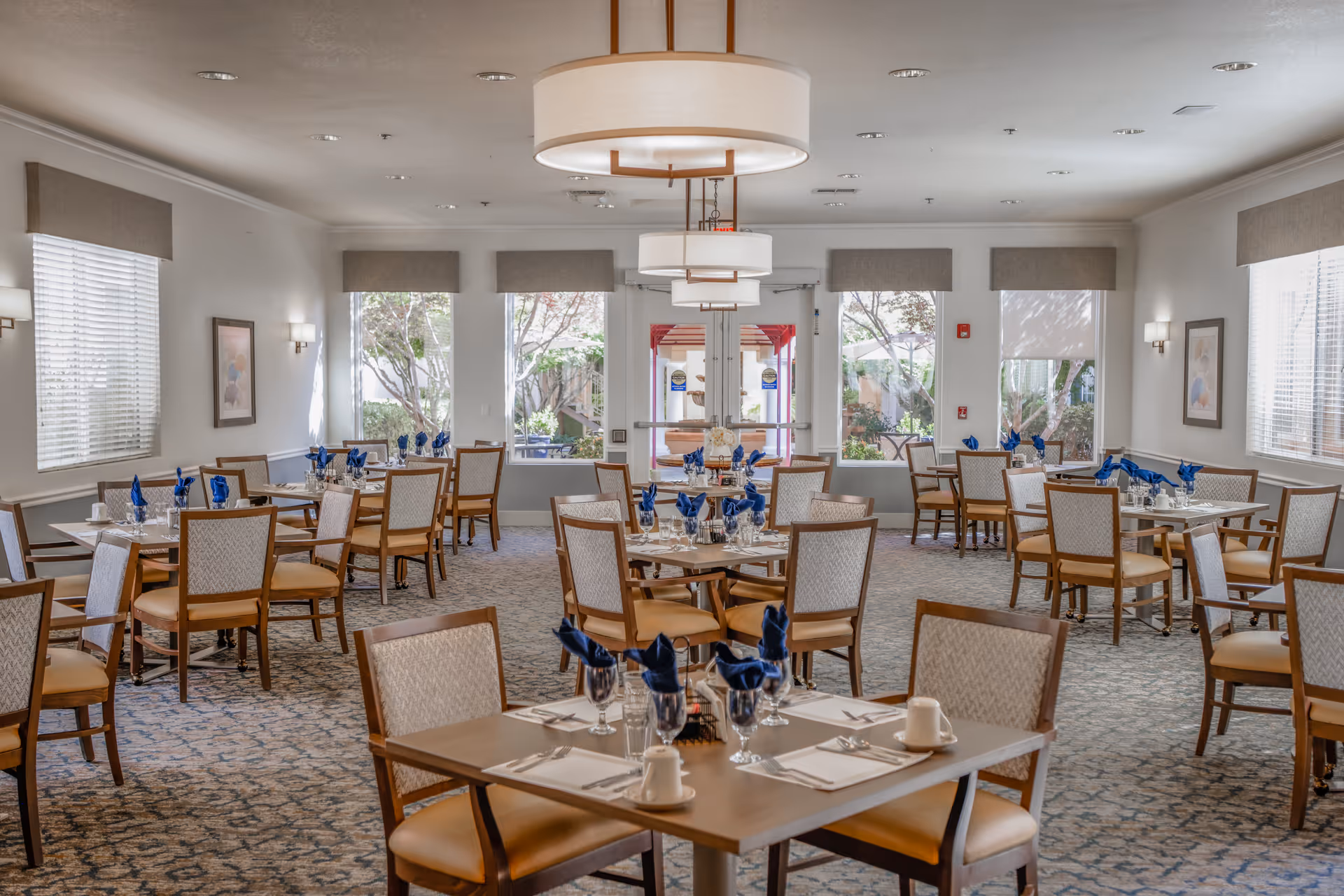 A bright and spacious dining room in a senior living facility with multiple tables set for meals. Each table has chairs with cushioned seats and backs, white napkins folded in a decorative manner, glassware, and silverware. Large windows allow natural light to fill the room, and there are modern light fixtures hanging from the ceiling. The walls are decorated with framed artwork and wall sconces.