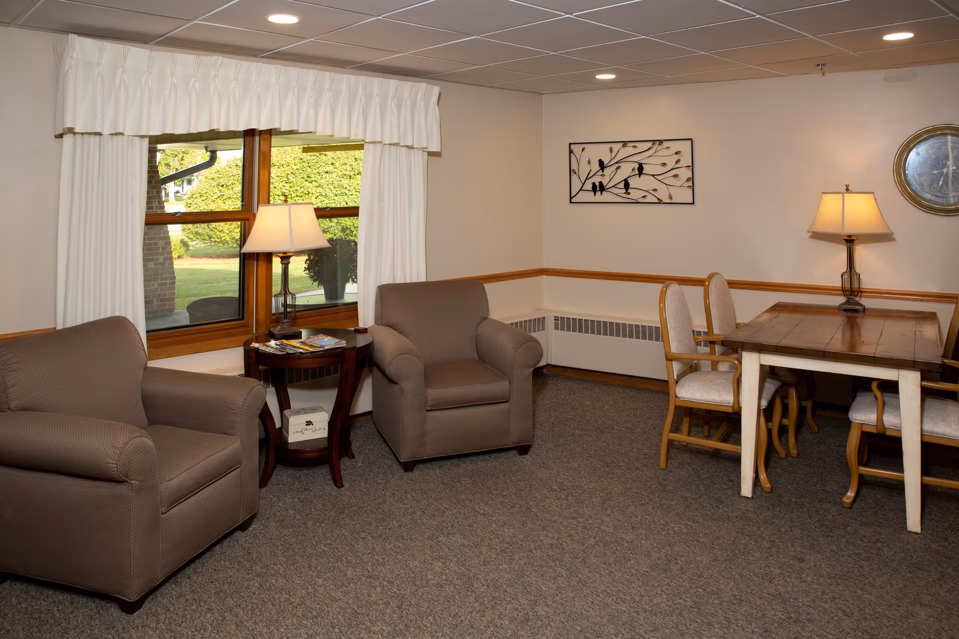 A cozy common area with two brown armchairs, a wooden side table with a lamp and magazines, a wooden dining table with four cushioned chairs, wall decorations including a metal bird artwork and a round clock, and a window with white curtains showing greenery outside.