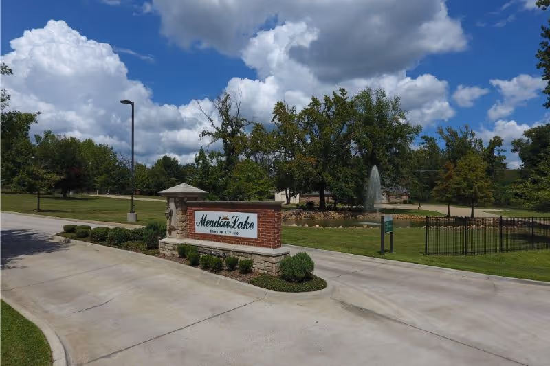 Entrance driveway to Meadow Lake Senior Living facility with a brick and stone sign displaying the facility name, surrounded by green grass, trees, and a water fountain in the background under a partly cloudy blue sky.