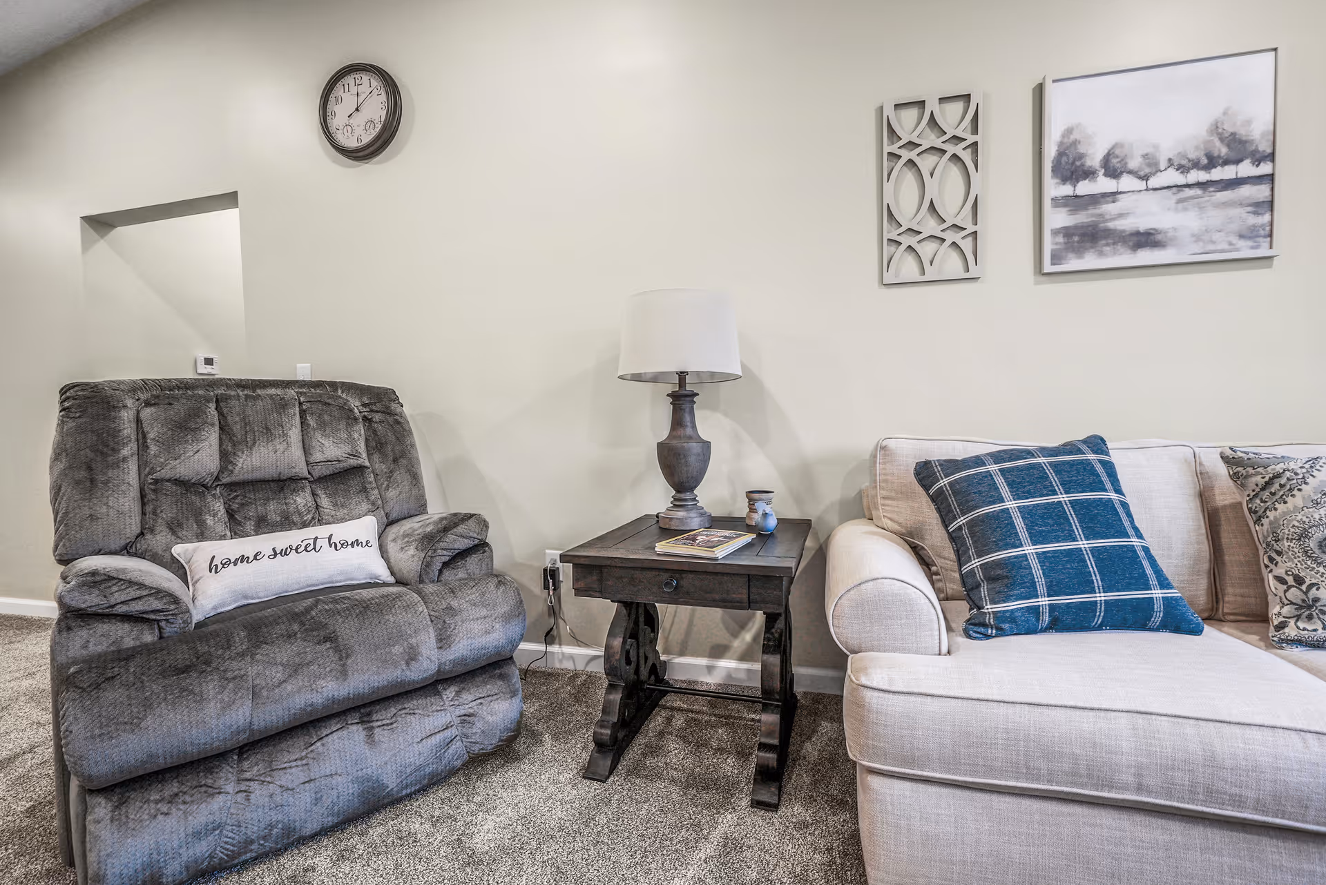 Cozy living room with a gray recliner and beige sofa flanking a wooden side table topped with a lamp and wall art above.