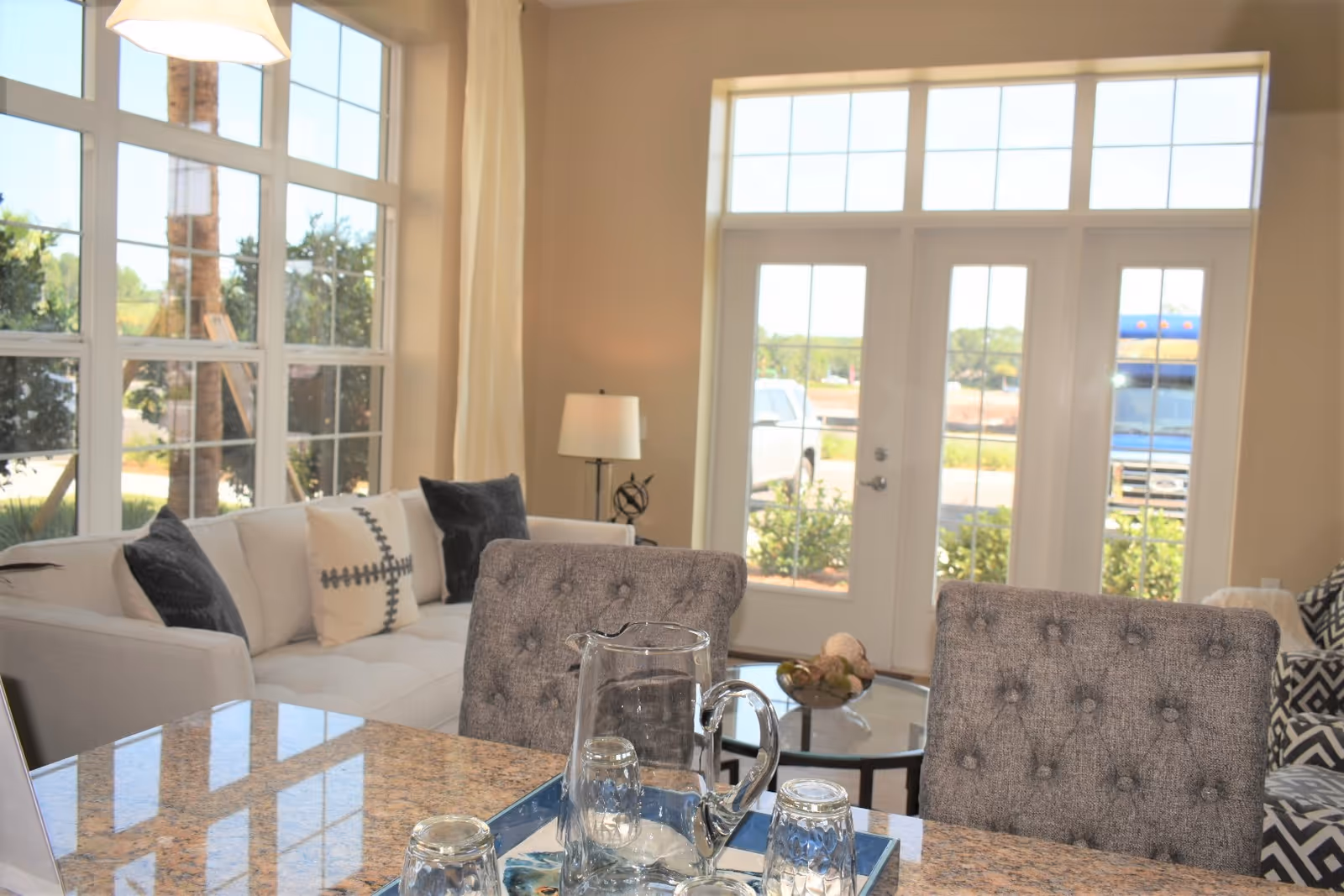 Bright open living area with tufted chairs and a sofa by large windows and French doors, seen from a granite island with a pitcher and glasses.