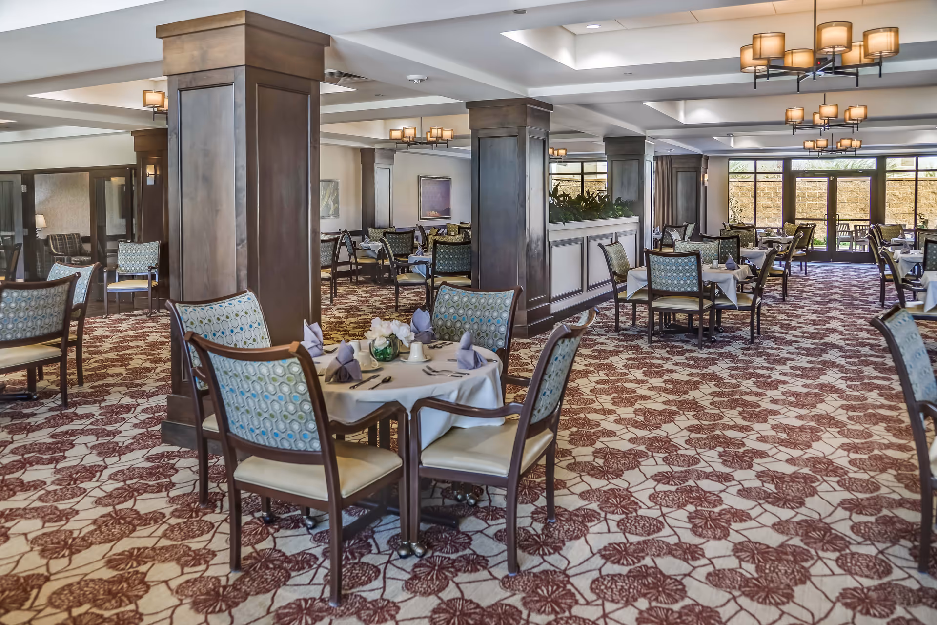 Spacious dining room with multiple tables covered in white tablecloths, each set with folded napkins, cups, and cutlery. The room features patterned carpet, wooden pillars, cushioned chairs with patterned backs, and large windows letting in natural light.