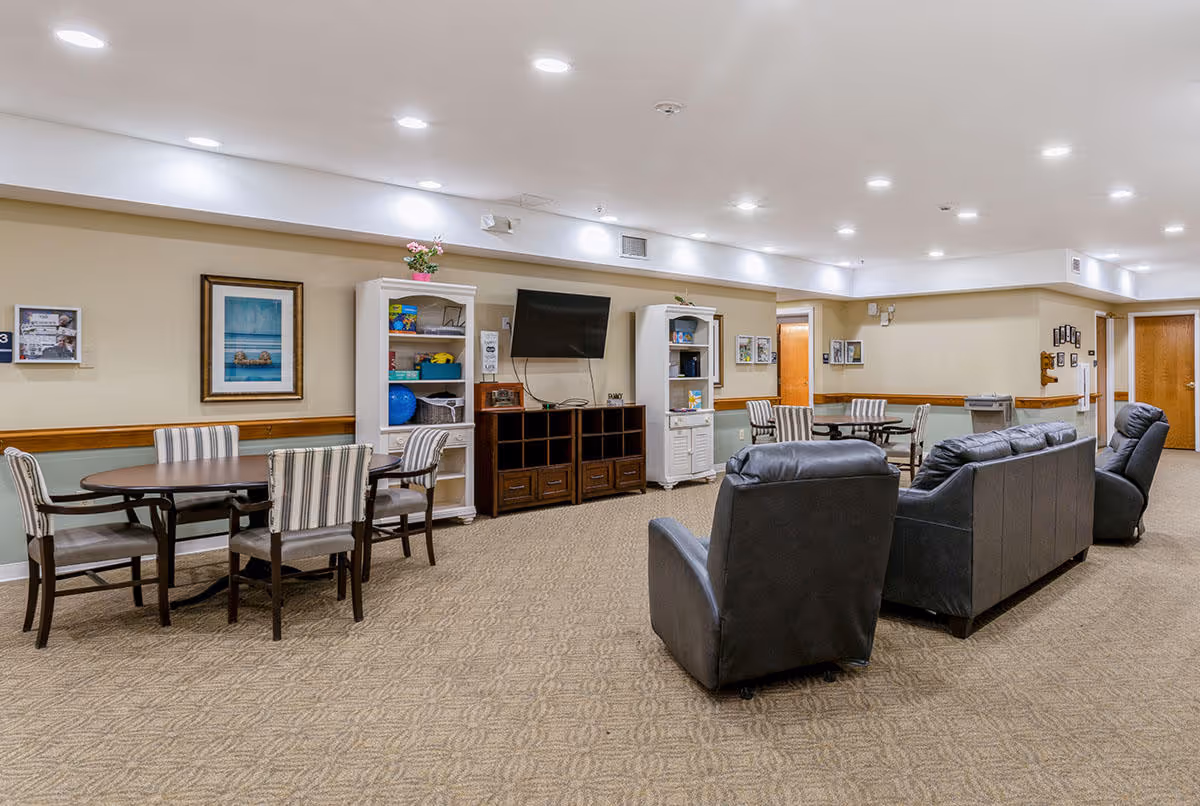 A spacious common area in a senior living facility featuring a seating arrangement with a black leather sofa and two matching armchairs facing a wall-mounted TV. There are two round tables with striped chairs along the wall, white shelving units with decorative items, and framed pictures on the beige walls. The room has carpeted flooring and recessed ceiling lights.